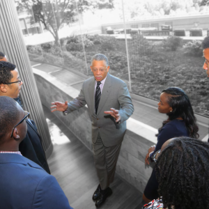 a group of people gather around one speaker in front of a wall of windows