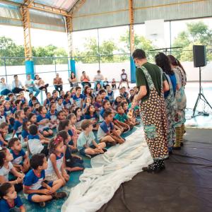 People in costumes stand in front of a large group of seated children in an open building.