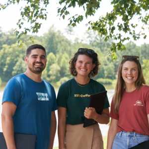 three smiling people in front of water and trees