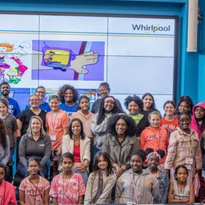 A group of children and adults posed in a classroom with a large digital screen behind them.