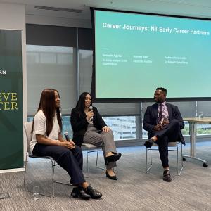 Three people with microphones seated at the front of a room. A projected display behind them.