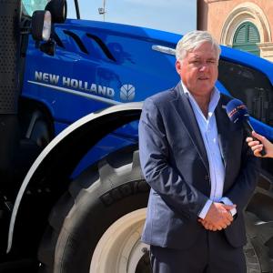 Carlo Lambro in front of a blue New Holland tractor, another holding a microphone for them. 