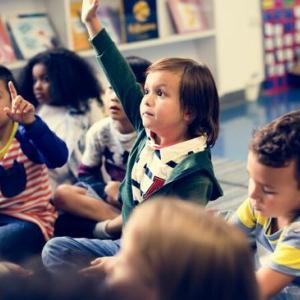 young children sitting on a classroom floor and raising their hands