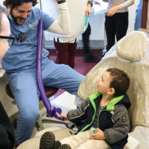 A dentist plays with a child in a dentist chair, a tooth mascot in the corner, others around smiling.