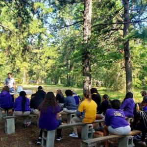 Students sitting on benches in a park setting watch a speaker