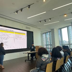 A classroom of students, two presenters at the front, a digital presentation behind them.