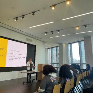 A classroom of students, a presenter at the front with digital display behind them.