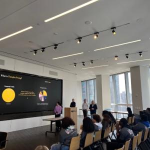 A classroom of students, multiple presenters at the front and digital display of pie charts behind them.