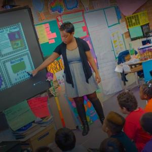 A teacher in front of a class of students, looking at a screen with lesson in area and perimeter on it.