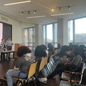 A classroom, three presenters at the front, a digital display behind them.