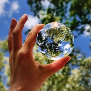 A hand holding a small clear globe. Trees and blue sky behind it.