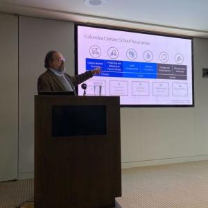 speaker at a podium gesturing to a graphic on a screen