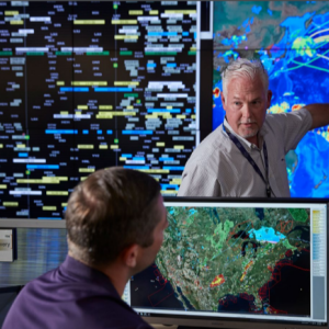 in a room with large screens on the walls, a person points at a global map while talking to a seated person looking at a computer screen with a map of the US