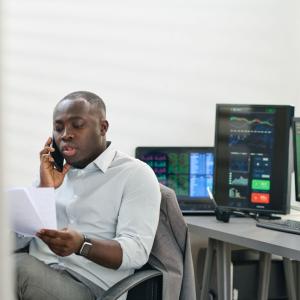 A person talking on a phone, looking at papers, their back to multiple computer screens.