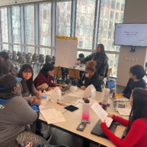 A group of people at a square table, papers strewn about. One at a paper board on an easel. 