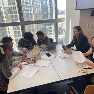 A group of people at a square table, papers strewn about. Large windows behind them.