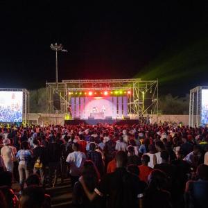 A large event with many people standing, looking at the stage and projected screens on each side. Performers on stage.