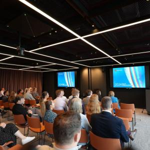 A seated group listening to a speaker at a podium at the front of the room.
