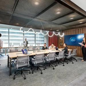 People seated at a long table with rolling office chairs, a long open wall, and person at a board near the front of the space.