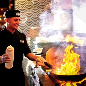 A chef smiling looking at a flaming wok on a stove.