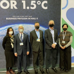 a group of five people in business attire, in front of a "For 1.5 degree C" banner. All are wearing medical masks.