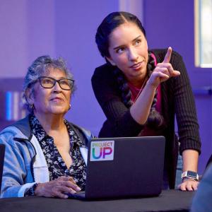 A person using a laptop with another person leant on the table next to them pointing to something in the distance 