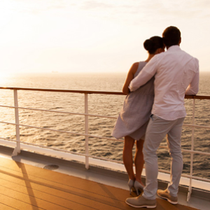 a couple hugging, leaning against the rail of a cruise ship. Setting sun in the background.