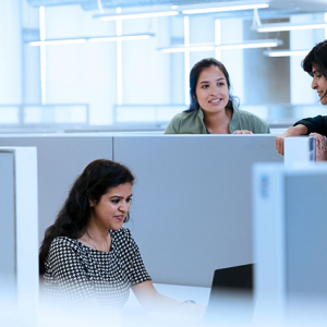 Two people standing, talking with a third who is seated at a desk in a cubicle, working on a laptop.