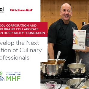 Two people standing behind a stove with pots of food. One holds a box for a kitchen aid pasta maker. "To Develop the next generation of culinary professionals. MHF and Whirlpool logos.