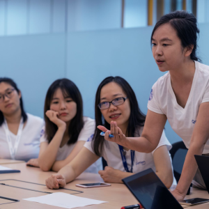 A group of people seated around a table, one standing and leaning towards another. 