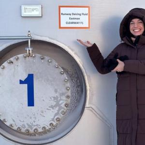 A smiling person pointing to a tank with a sign "Runway deicing fluid. Eastman."
