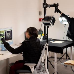 Two people in a room, one at a computer desk, pointing to an image on the screen. The other adjusting equipment above a photograph on a table.