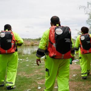 Three people seen from the back walking away, in high-vis suits with Direct Relief backpacks