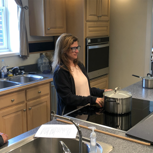 woman wearing frosted glasses stands at an induction range and touches a silver pot with the back of her hand