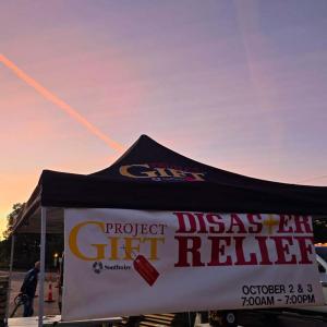 A pop-up canopy with Project Gift banner hanging on the front, Outside in a parking lot. A setting sun in the background.
