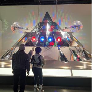 A shiny silver metal triangular spaceship with red and blue lights and eight metal legs is displayed in National Museum of African American History and Culture. Two museum visitors stand in front of the spaceship and read a description panel.