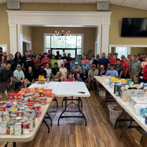 A large group of people posed behind tables full of canned food items and supplies.