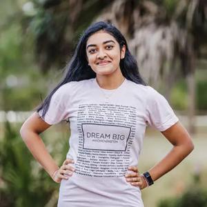 A person smiling at the camera, hands on their hips. Wearing a "dream big" t-shirt. 
