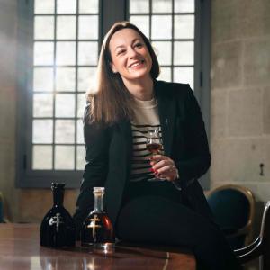 Agathe Boinot half-seated on a table. Bottles of cognac next to her. A small glass in her hand.