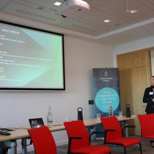 A person speaking at the front of a room with a large display behind them.