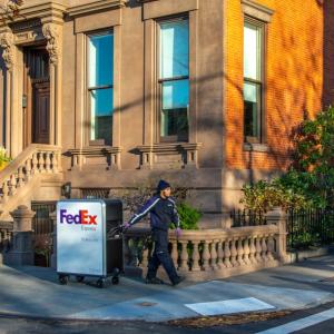 A person in FedEx uniform pulling a tall cart with FedEx Express logo on it going around a corner on the sidewalk in front of a house.