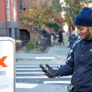 A person in FedEx uniform next to a tall cart with FedEx Express logo on it. They are looking at a hand-held device on a sidewalk in a residential area.