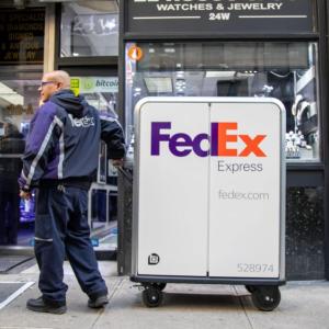 A person in FedEx uniform pulling a tall cart with FedEx Express logo on it, waiting outside of a jewelry store.
