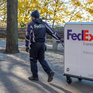 A person in FedEx uniform pulling a tall cart with FedEx Express logo on it through a park setting.