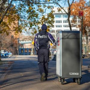 A person in FedEx uniform pulling a tall cart with FedEx Express logo on it down a sidewalk as a car goes by.
