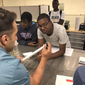 Three students watch an instructor assemble a piece of equipment