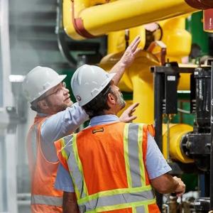 Two people looking at large yellow indoor piping, each wearing hard hats and high-vis vests.
