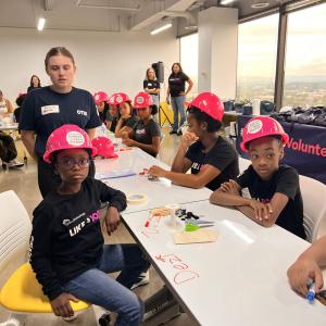 Children seated at long table wearing matching pink hard hats.
