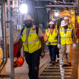 Group of five people on a marked walkway, each wearing high-vis vests, gloves and hard hats in an industrial setting