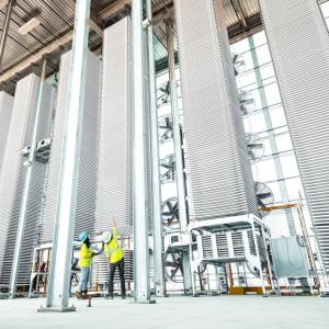 Two people in high-vis vests looking at a tall stack of metal sheets in a row of other stacks.